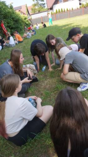 Młodzież siedzi w kole na trawie. W tle inna grupa.