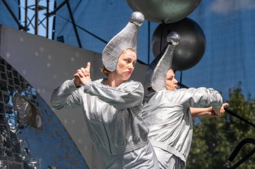 Dwie kobiety w srebrnych, kosmicznych strojach z dziwnymi nakryciami głowy składają ręce w geście strzału.