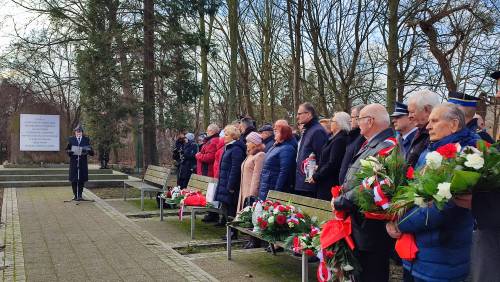 Widok na zebranych gości stojących za ławkami. W rękach trzymają kwiaty. W tle pomnik i przemawiająca osoba.