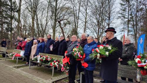 Widok na zebranych gości stojących za ławkami. W rękach trzymają kwiaty.