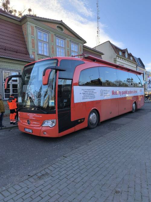 Czerwony autobus - mobilny punkt krwiodawstwa stoi na parkingu przed zabytkowym budynkiem.