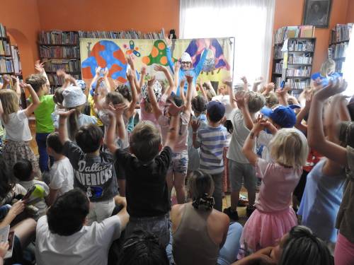 Przedstawienie w bibliotece, dzieci stoją z rękami w górze, przed nimi Smerf