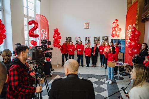Uroczystość, czerwone stroje, balony, przemówienie z kamerą w kadrze.