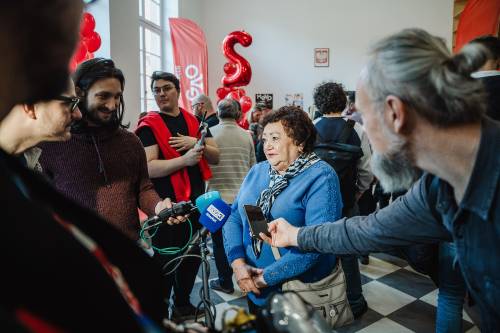 Kobieta w niebieskim swetrze i szaliku udziela wywiadu, otoczona przez kilku mężczyzn z mikrofonami.