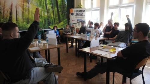 Uczniowie siedzą przy stolikach i podnoszą ręce głosując