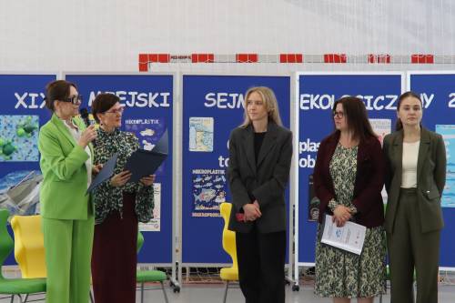 Nauczyciele i dyrektor szkoły stoją przed tablicą z napisem XIX Miejski Sejmik Ekologiczny