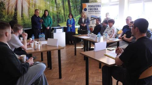 Młodzież siedzi przy kilku stolikach słuchając prowadzących spotkanie
