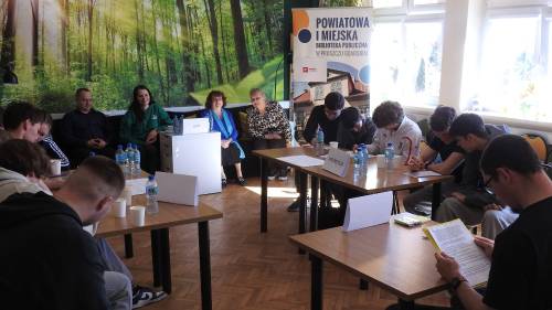 Grupki uczniów siedzą przy stolikach i czytają to co jest napisane na kartkach