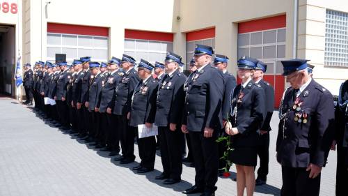 Strażacy stoją w szeregu na placu przed budynkiem