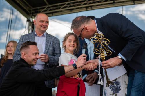 Burmistrz Pruszcza przekazuje dziewczynce duży klucz, inny mężczyzna klęczy z mikrofonem