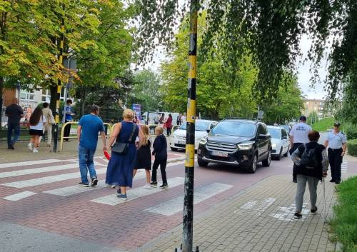 Strażnik miejski stoi na chodniku przy przejściu dla pieszych, w pobliżu którego przechodzą dzieci.