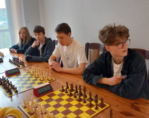 Młodzież siedząca przy stole z rozłozonymi szachownicą i szachami, czekają na rozpoczęcie gry.
