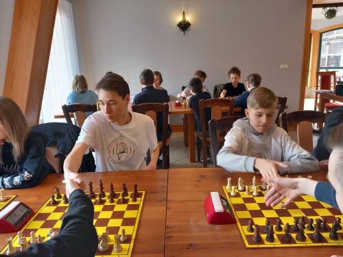 Młodzież siedząca przy stole z rozłozonymi szachownicą i szachami, rozpoczynają grę podając sobie rekę.